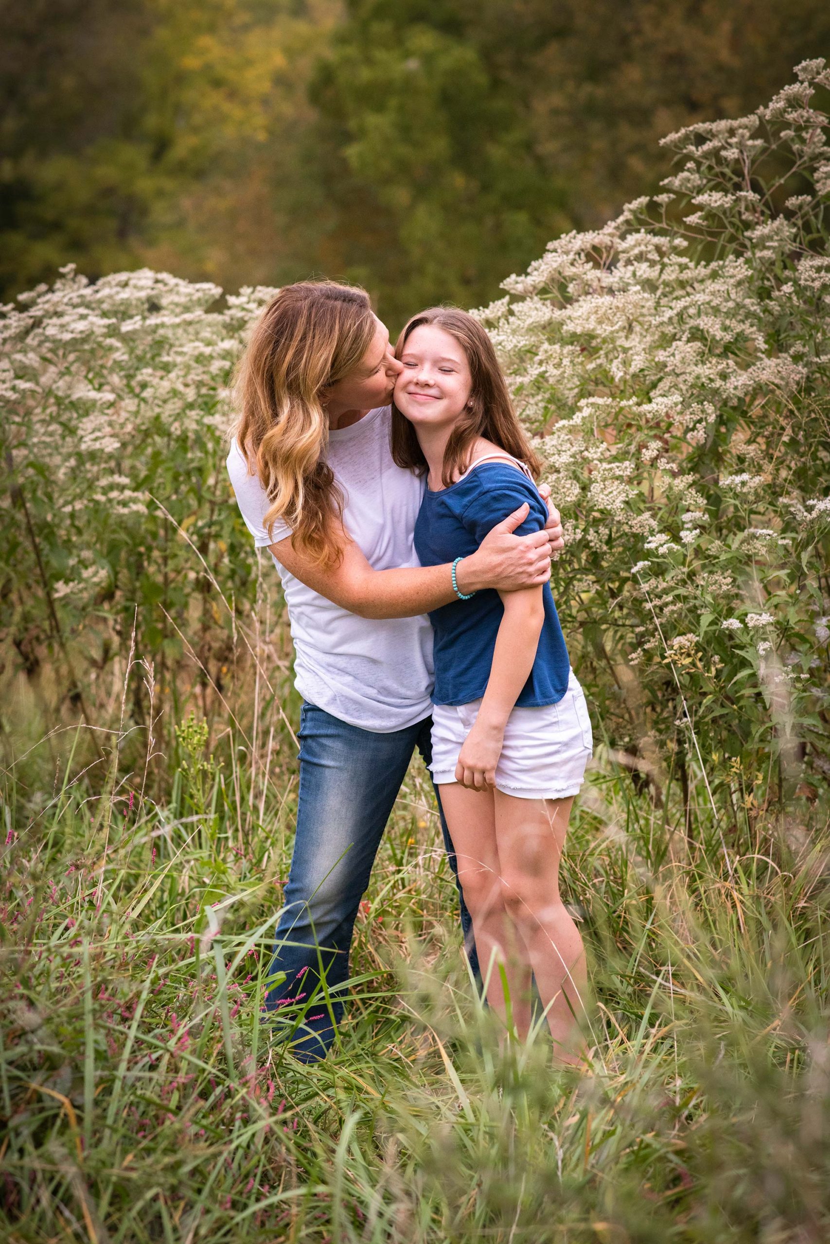 Mom gives tween daughter a kiss on the cheek in a field of flowers.