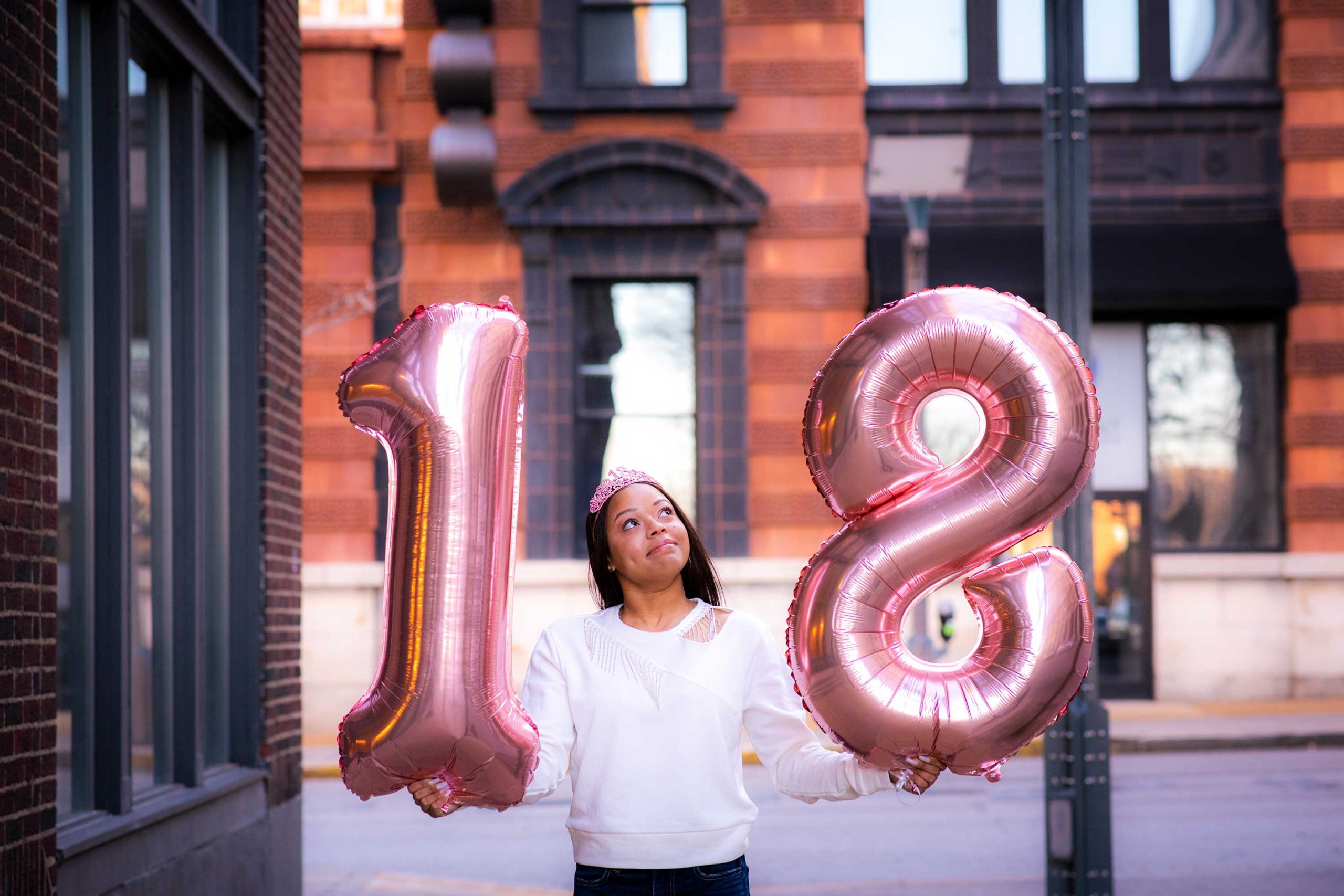 Girl in an urban setting holding up large number balloons that say "18" in celebration of her 18th birthday.
