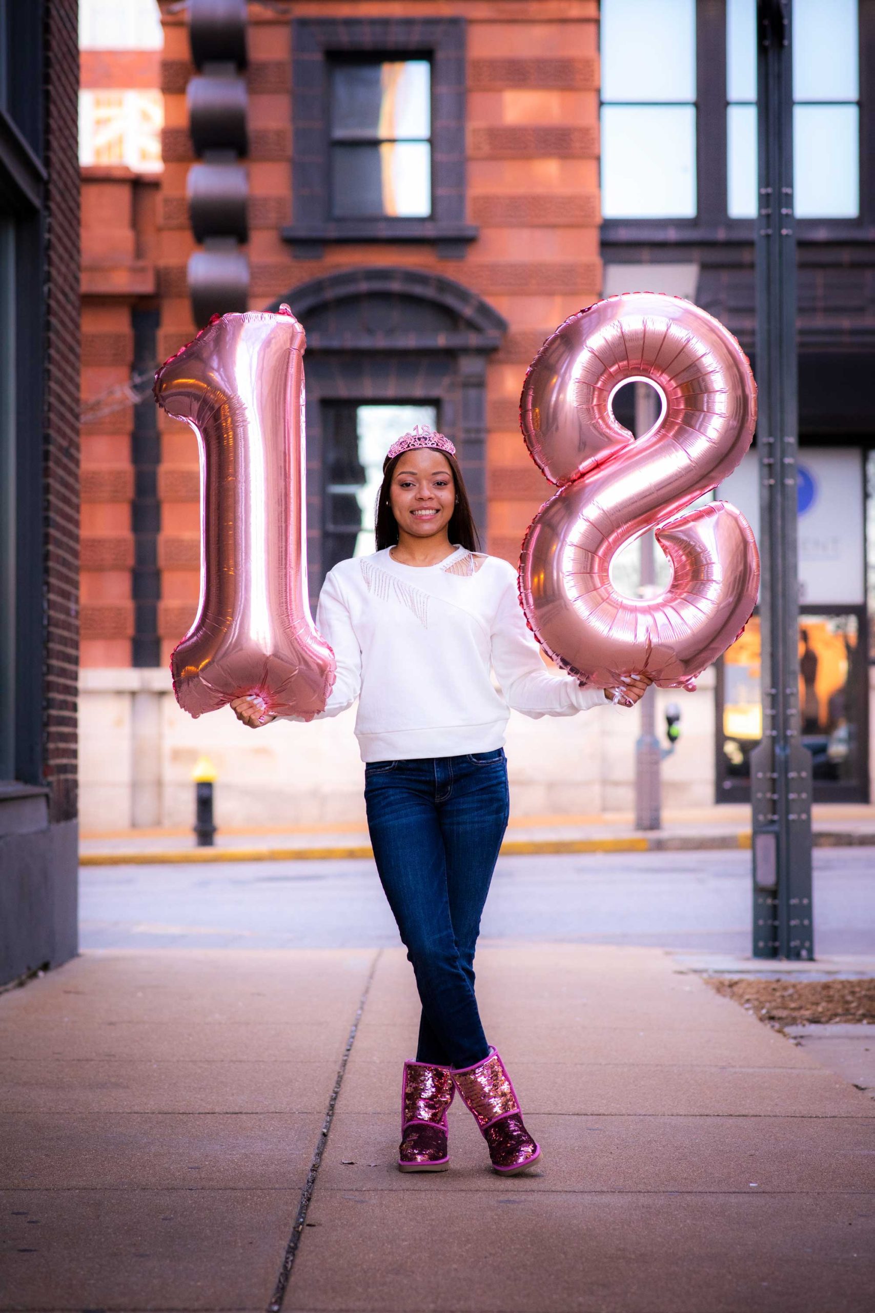 Girl in an urban setting holding up large number balloons that say "18" in celebration of her 18th birthday.