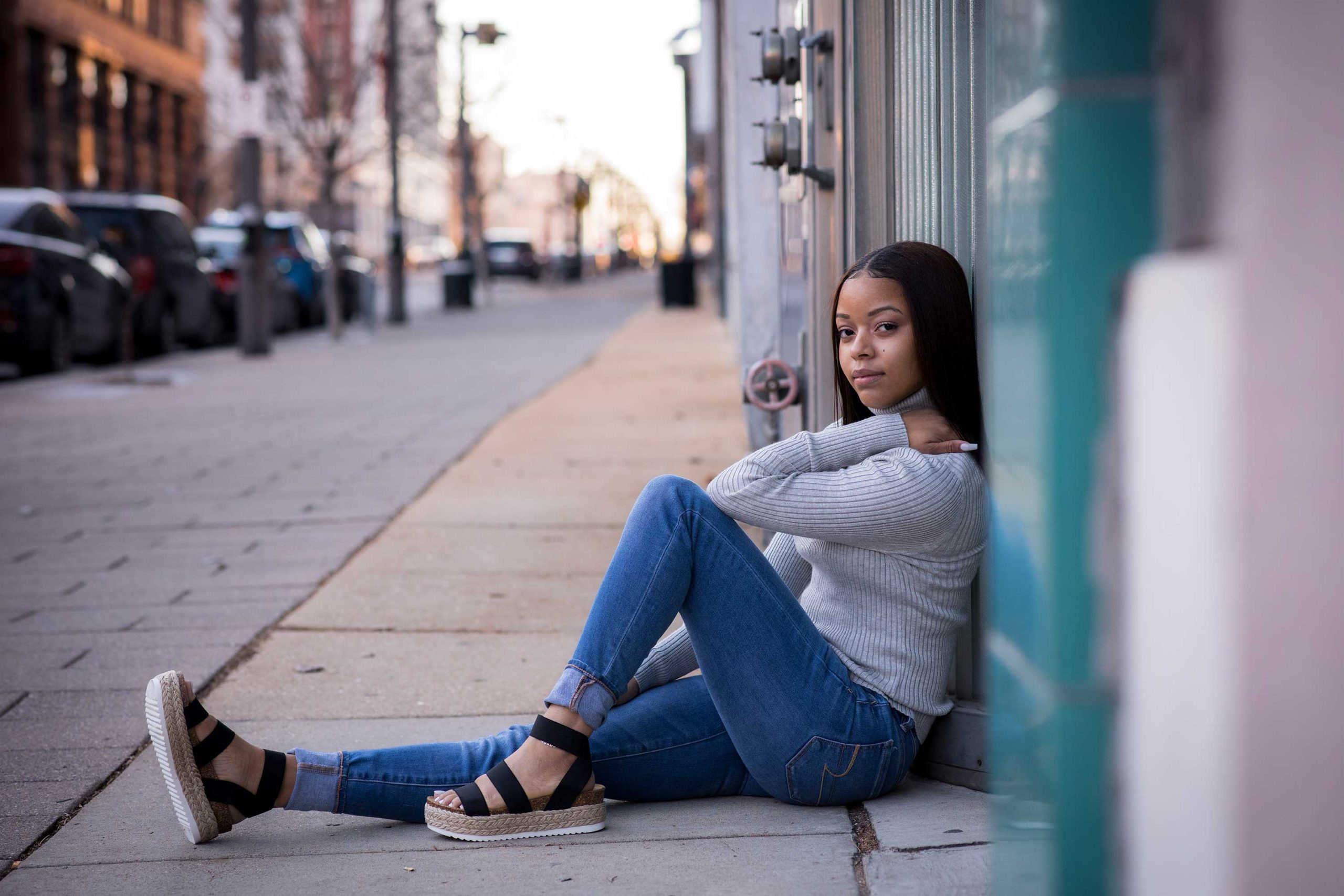 Girl in jeans and sandals in an urban setting sitting against turquoise and silver building.