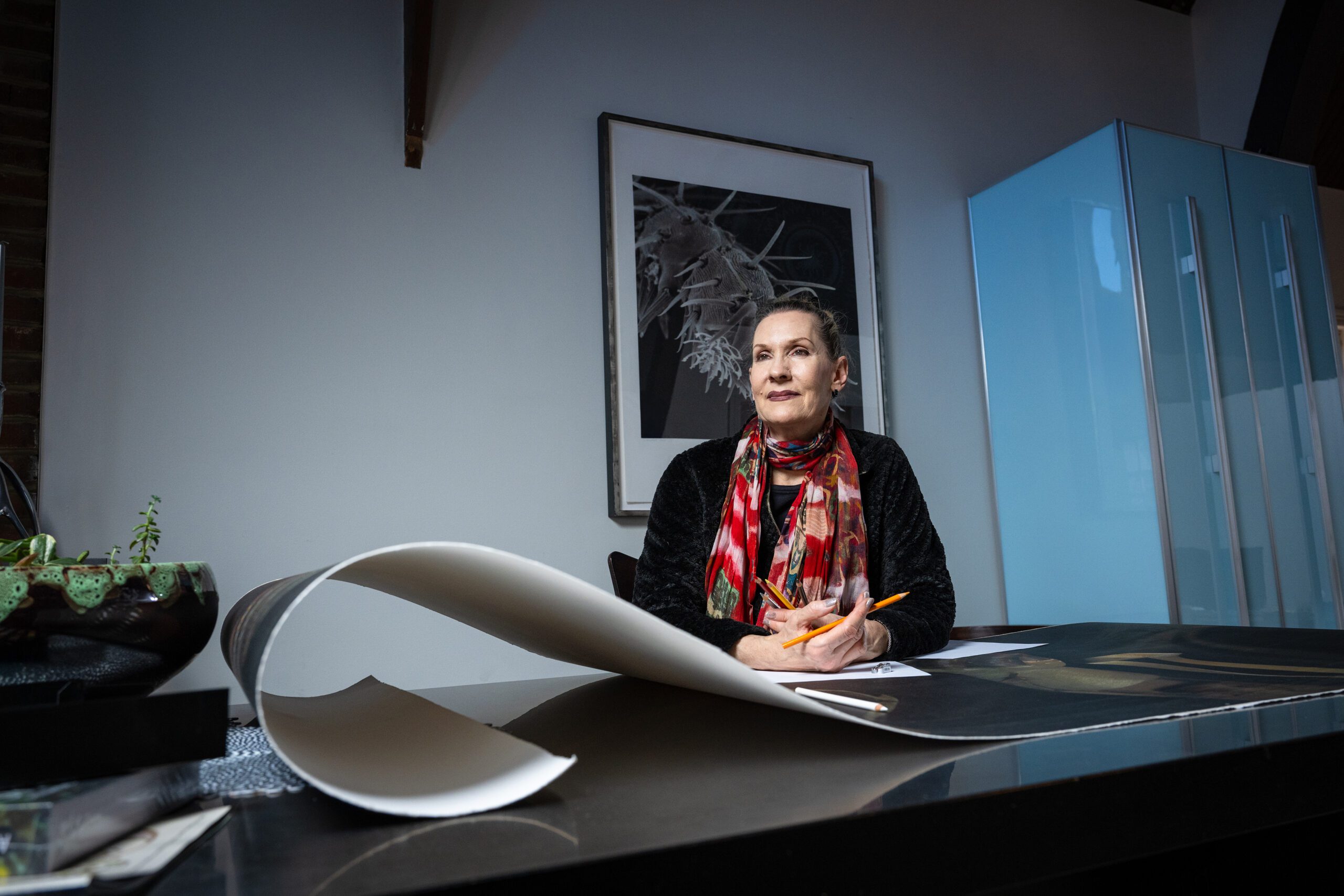 Washington University Faculty and Artist Patricia Olynyk poses for a portrait in her home with one of her photos on the wall and on the table in front of her.