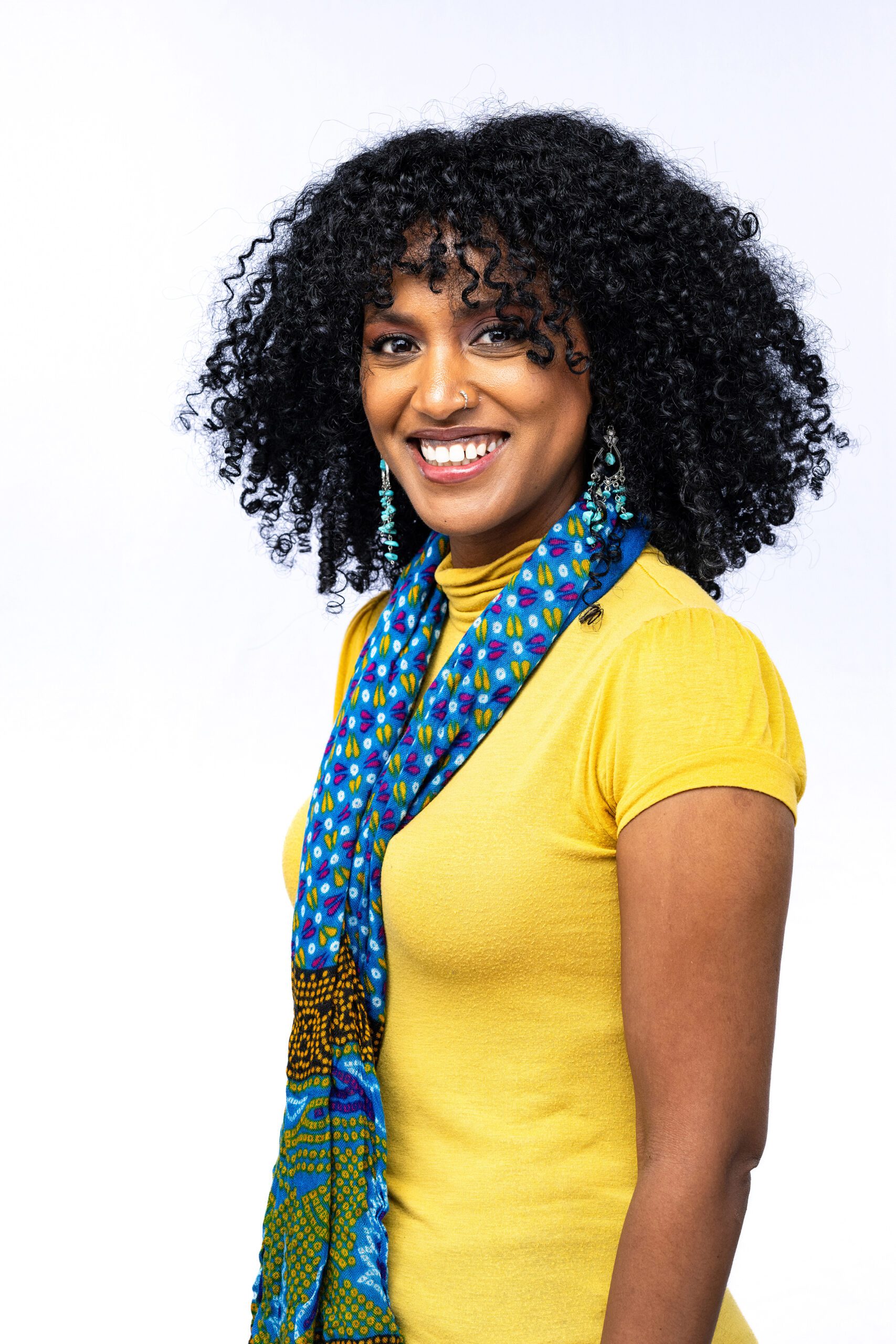 Headshot of a female in a yellow shirt with a blue scarf with a white backdrop.